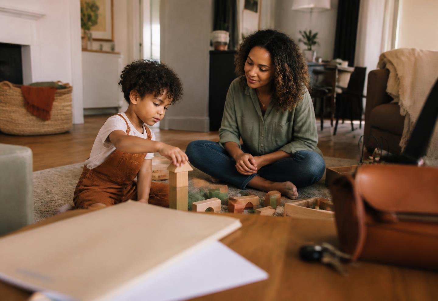 A mother sits on the floor with her young child, who is building with wooden blocks, while a closed appointment folder and car keys rest on the table nearby.