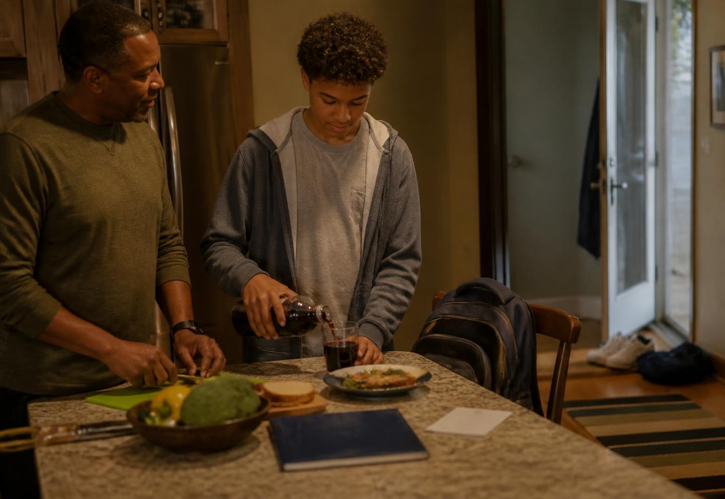 A father prepares dinner at the kitchen counter while his teenage son, just home from school, stands beside him making a snack, with a backpack on a chair and a closed school folder on the counter.
