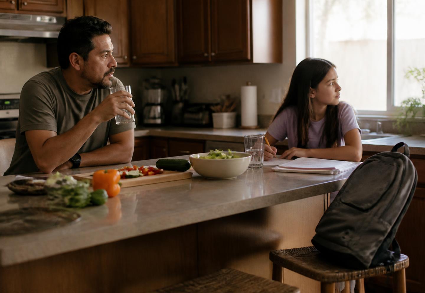 A father and his tween daughter share a kitchen island in the early evening after a frustrating moment — he holds a glass of water, she sits sideways with a pencil looking toward the window, both quiet and still connected.